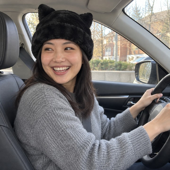 Illustrative photo of a woman in a Black Cleo cat ear hat and a gray sweater, hands on the steering wheel.