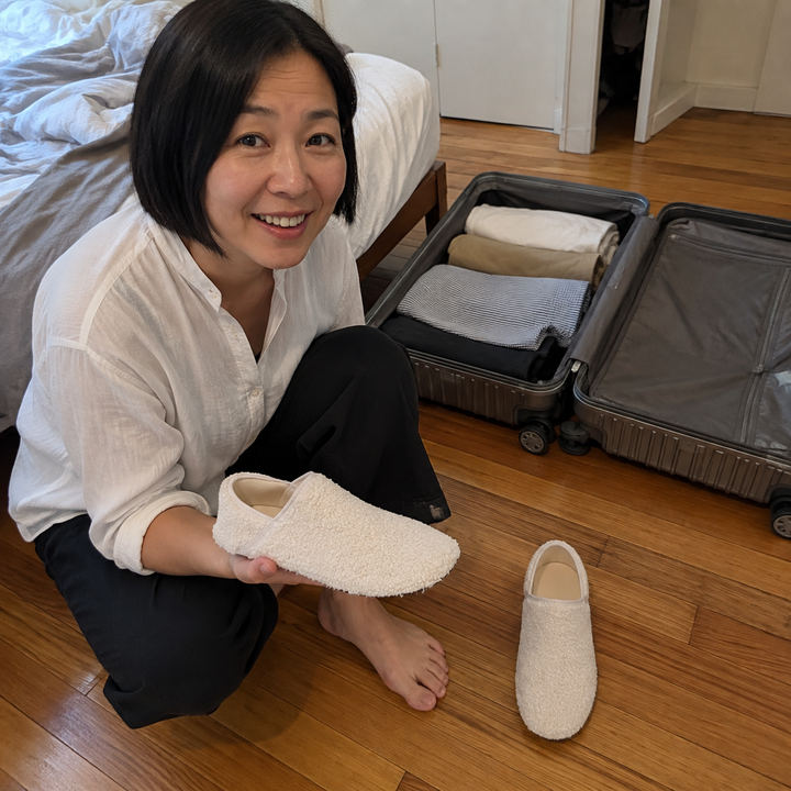 A woman crouched by an open case on the floor, one barefoot travel slipper sock resting flat on her upturned palm and the other standing on the floor beside her