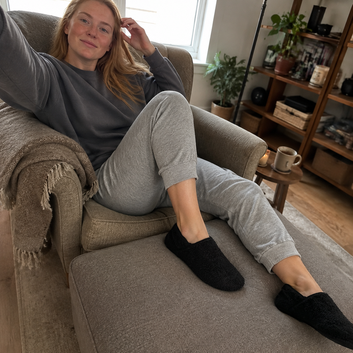 A woman with her feet up on an armchair, one knee drawn up and one leg out, wearing barefoot travel slipper socks
