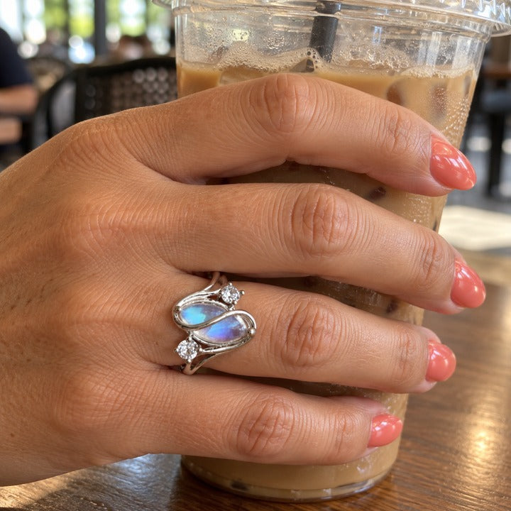 Hand around an iced coffee at a café table, wearing the simulated moonstone ring