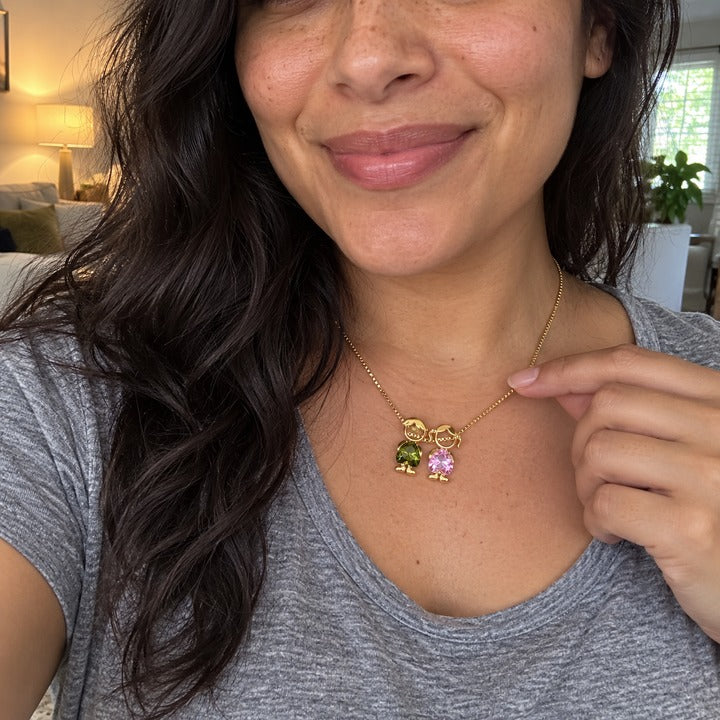 Selfie at home, a soft smile, her fingertip by the gold-tone necklace's boy and girl charms in olive green and pale pink