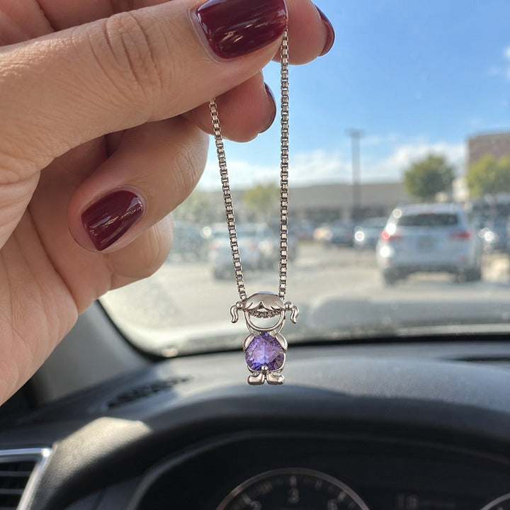 The silver-tone necklace with one girl charm and a violet heart-cut stone, dangling from her fingers in a parked car