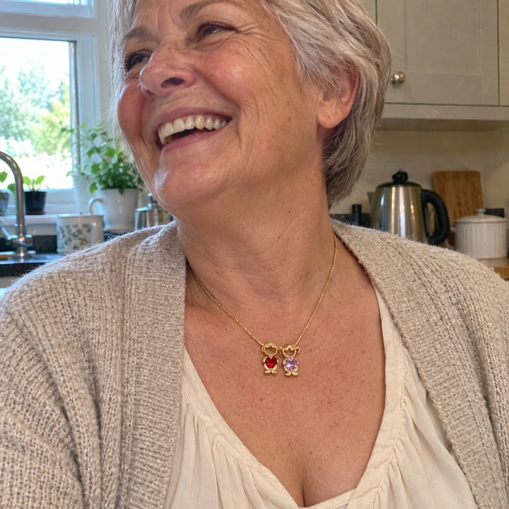 A grandmother laughing at the kitchen table in the gold-tone necklace, a boy and a girl charm in bright red and pale lavender