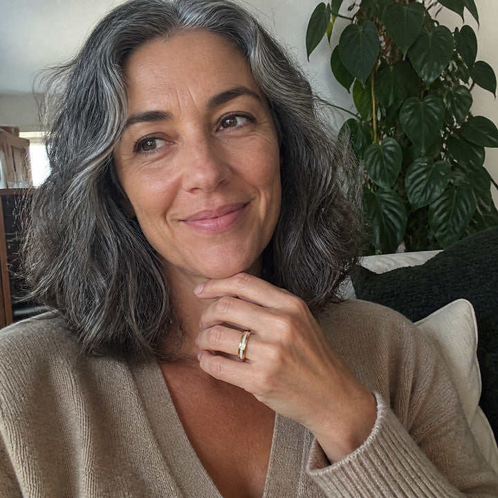 A woman on a sofa beside a houseplant, wearing the Gold Crystal Spinner Anxiety Ring