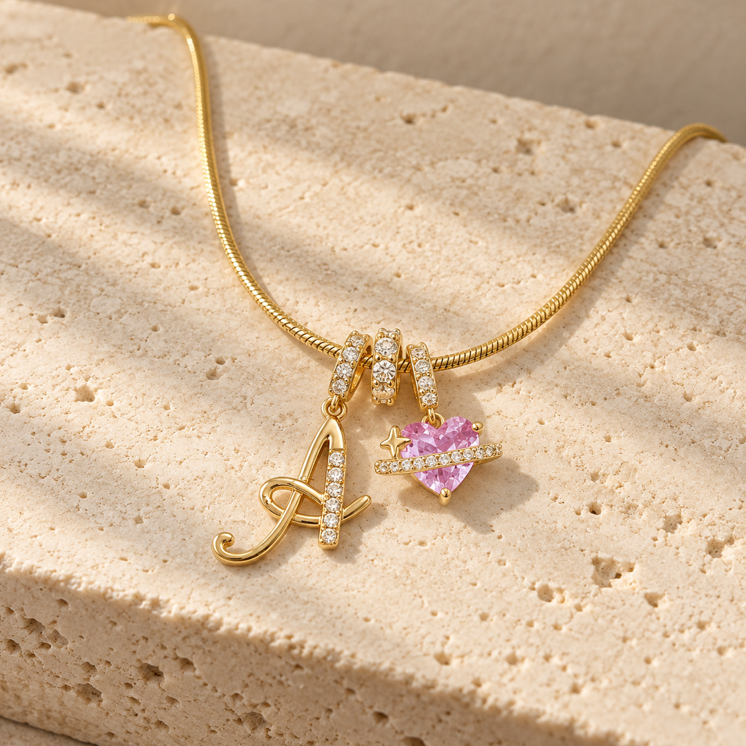 Cherie initial necklace in gold, script initial A beside a pink heart charm, laid across a travertine block