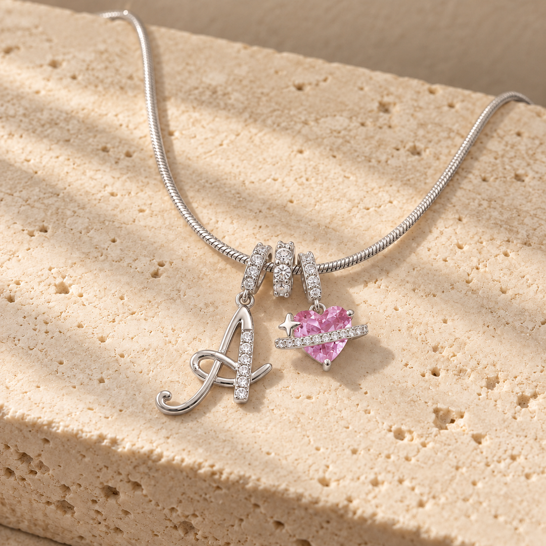 Cherie initial necklace in silver, script initial A beside a pink heart charm, laid across a travertine block