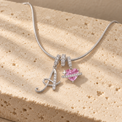 Cherie initial necklace in silver, script initial A beside a pink heart charm, laid across a travertine block