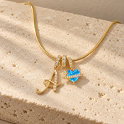 Cherie initial necklace in gold, script initial A beside a blue heart charm, laid across a travertine block