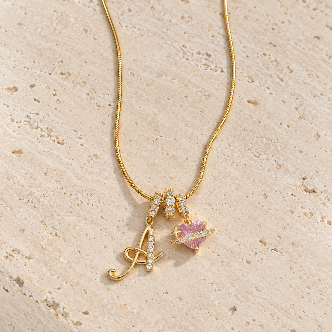 The gold Cherie initial necklace with initial A, its three stone-set rings and the pink heart on travertine