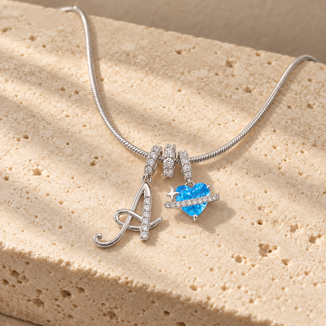 Cherie initial necklace in silver, script initial A beside a blue heart charm, laid across a travertine block