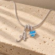 Cherie initial necklace in silver, script initial A beside a blue heart charm, laid across a travertine block