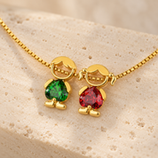 Close-up of a boy and a girl charm on a gold-tone box chain, green and deep red heart-cut stones, the girl with pigtails