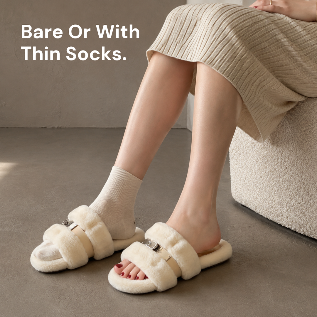 A woman seated in cream fur slippers, wearing one fine-gauge cream sock while the other foot is bare