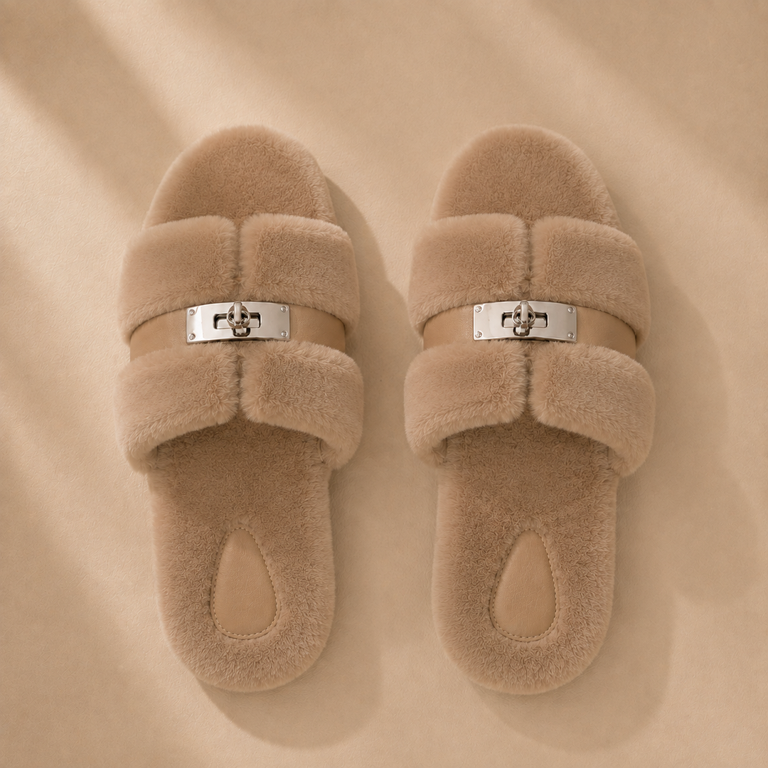 Top view of a pair of taupe fur slippers, the metal buckles centred on the strap between the two fur bands
