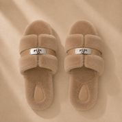 Top view of a pair of taupe fur slippers, the metal buckles centred on the strap between the two fur bands
