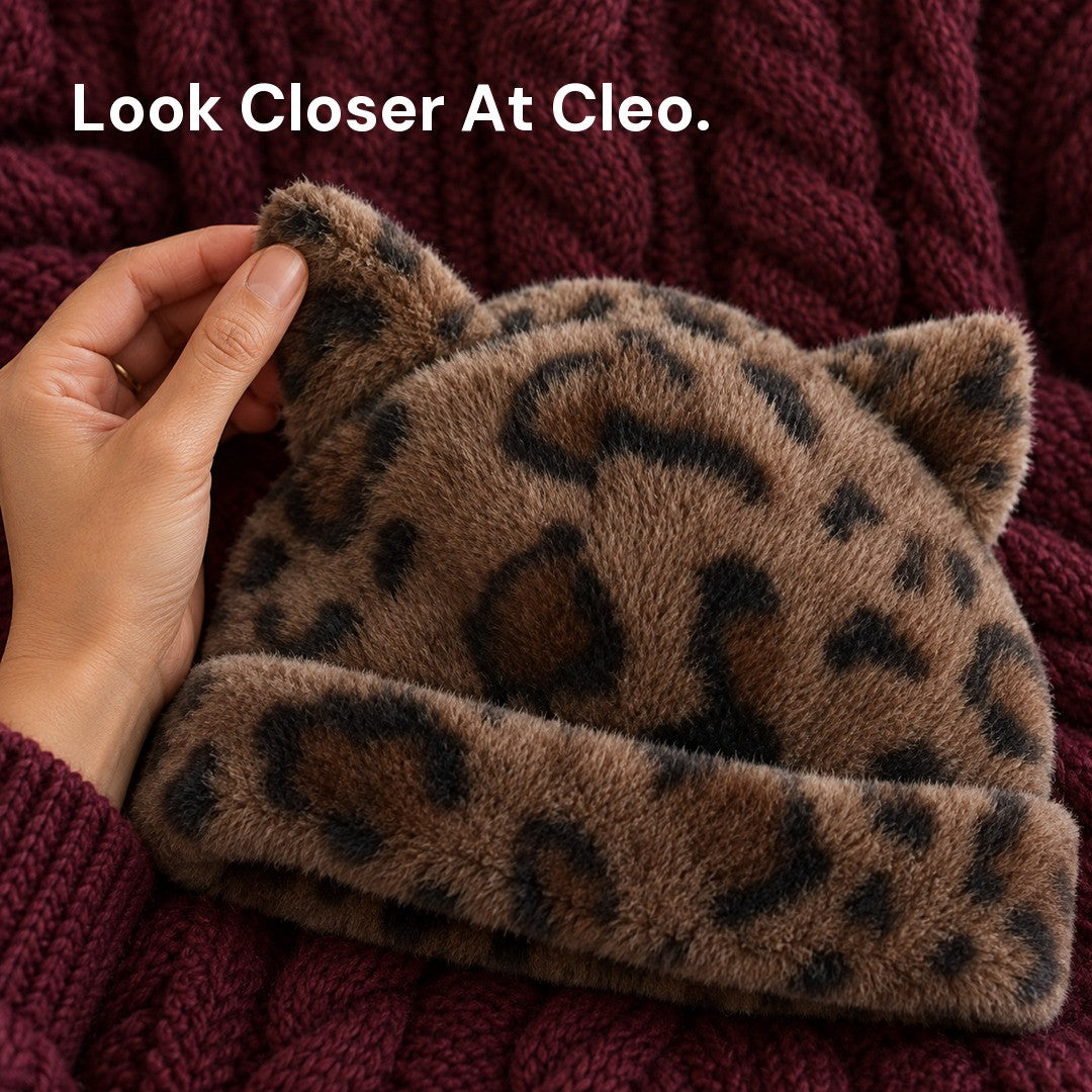 A hand lifting one plush ear of the Leopard Cleo cat ear hat laid on a burgundy cable-knit sweater.