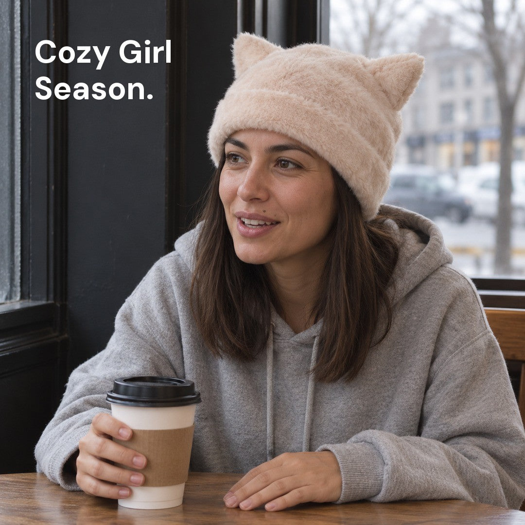 Woman in an Apricot Cleo cat ear hat and a gray hoodie holding a coffee cup at a café table.