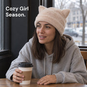 Woman in an Apricot Cleo cat ear hat and a gray hoodie holding a coffee cup at a café table.