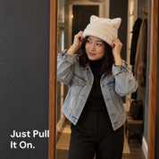 Woman in a denim jacket pulling on a White Cleo cat ear hat in a hallway mirror.