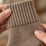 Two hands holding the folded cuff of a mid-calf sock at close range, the chunky rib of the cuff above the fold and the finer knit of the shaft below it