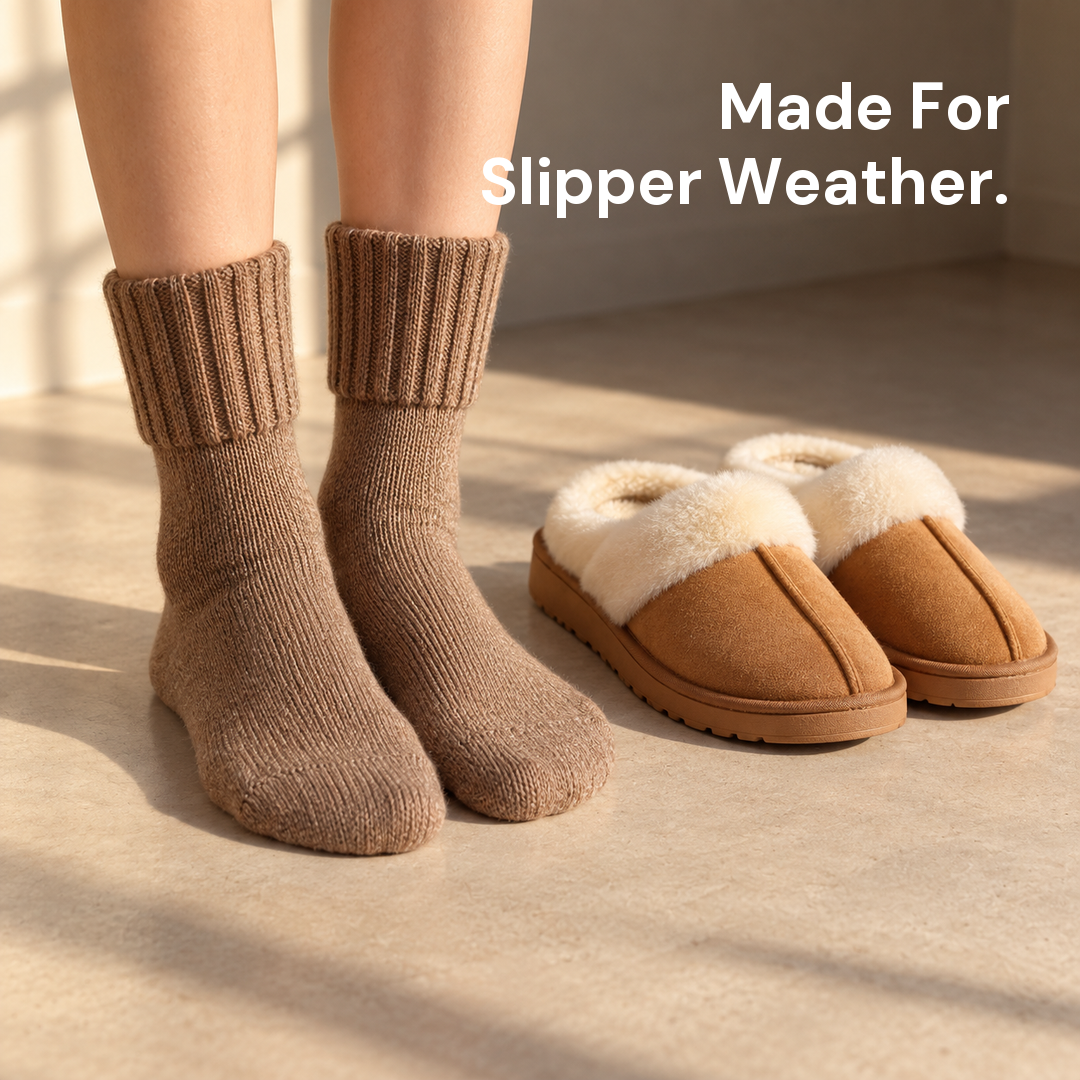 Feet in ribbed mid-calf socks resting on a floor beside a pair of slippers in a bright room