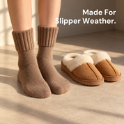 Feet in ribbed mid-calf socks resting on a floor beside a pair of slippers in a bright room