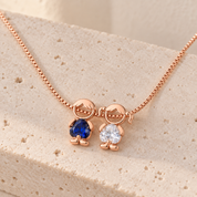 Rose gold-tone necklace on travertine with a boy and a girl charm, a royal blue and a clear heart-cut stone