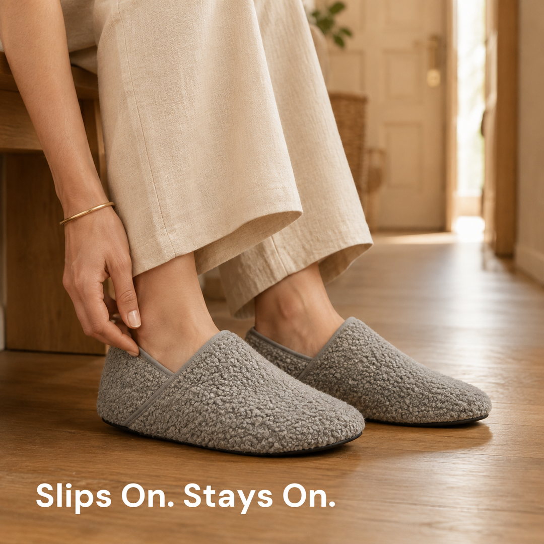 Both feet in grey slipper socks on a wood floor, one hand closed on the near shoe's bound collar
