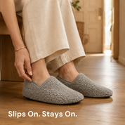 Both feet in grey slipper socks on a wood floor, one hand closed on the near shoe's bound collar