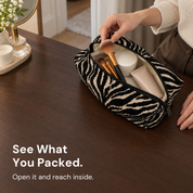 A hand reaches for a makeup brush inside an open black and cream zebra pouch on a wooden table.