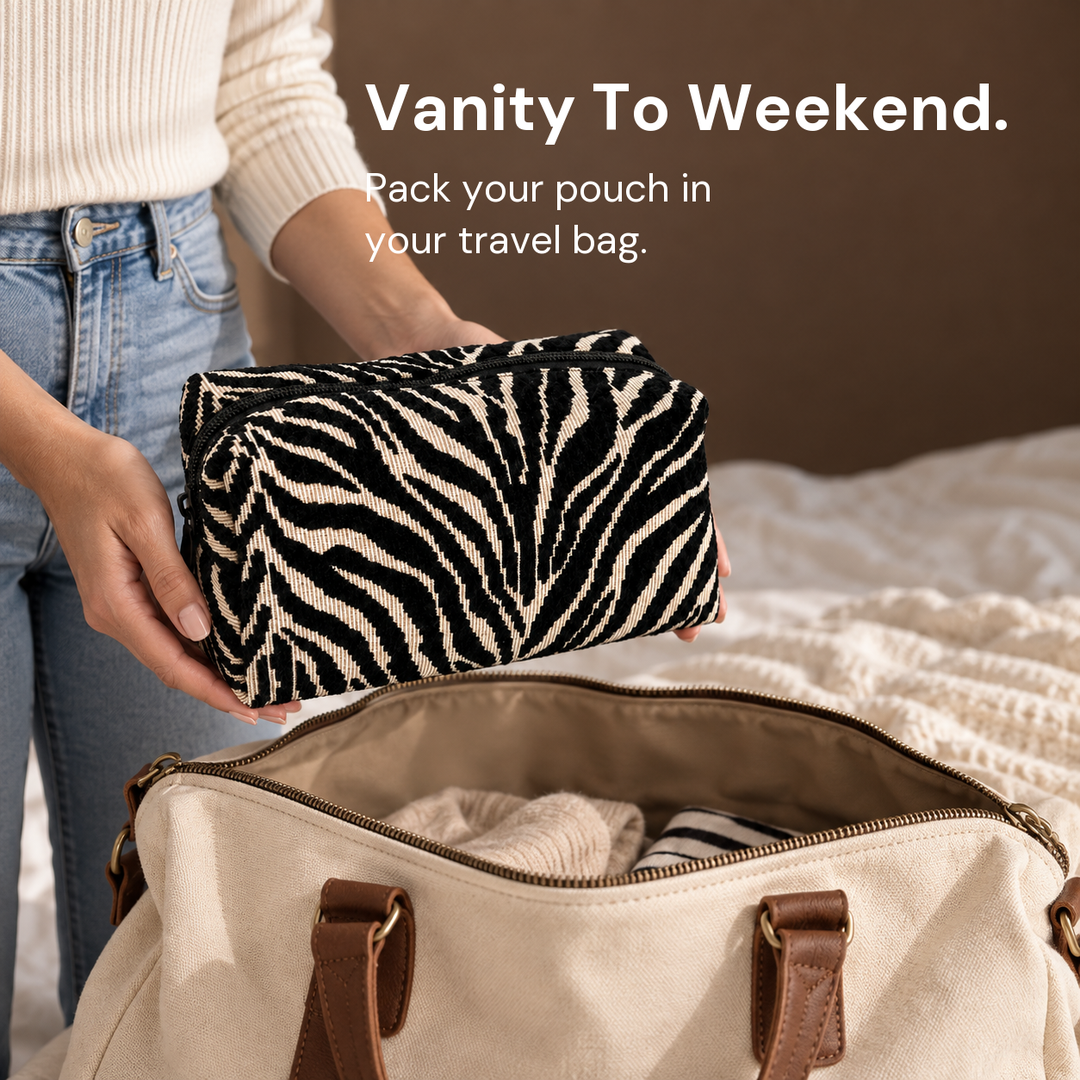 Hands hold a closed black and cream zebra pouch above an open cream travel bag on a bed.