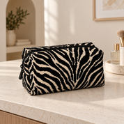 Black zebra makeup pouch standing closed on a cream vanity beside a tray of cosmetics.