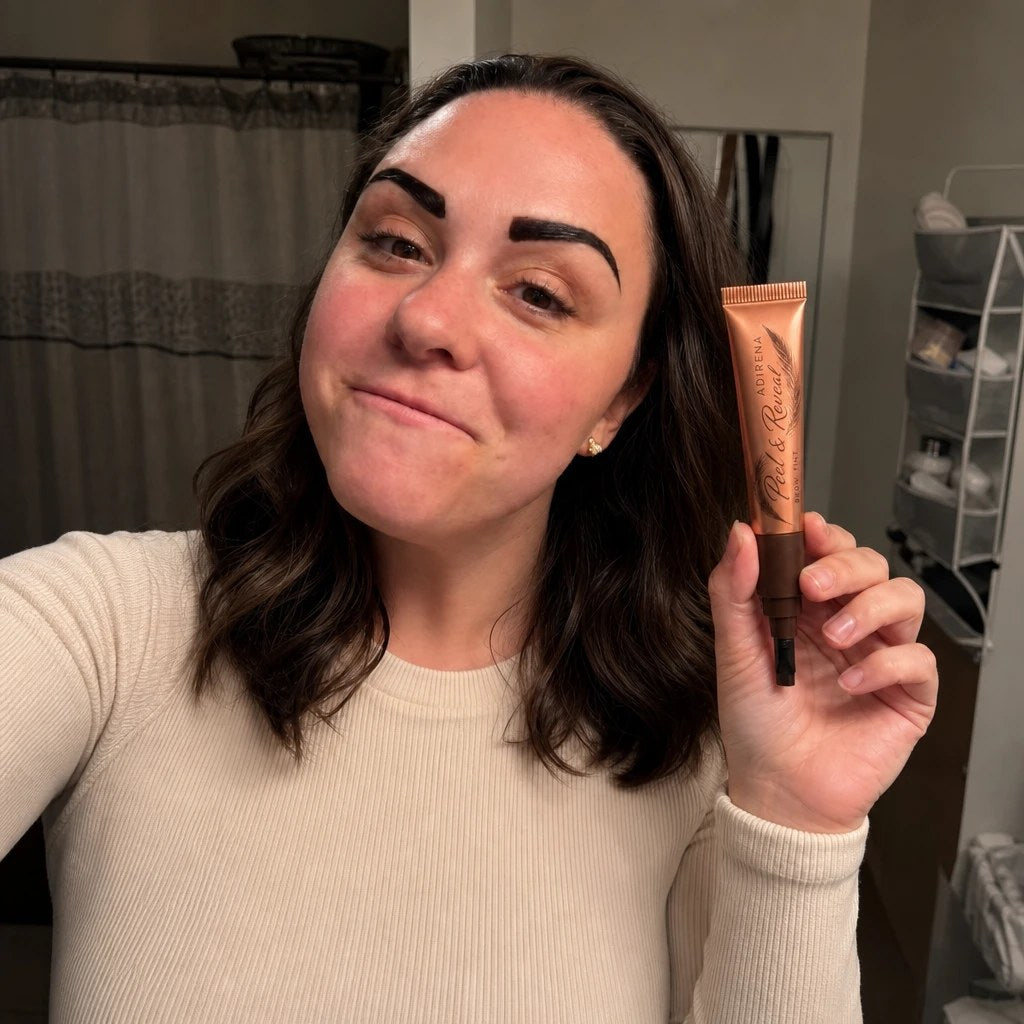 The reviewer in her bathroom holding the Adirena Peel & Reveal Brow Tint tube