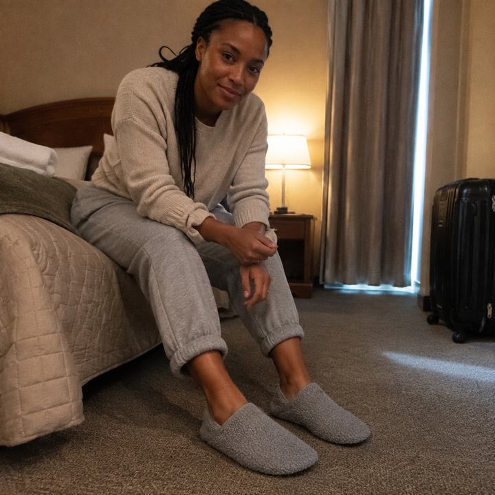 A woman sitting on the edge of a hotel bed, shoes on, feet flat, wearing barefoot travel slipper socks