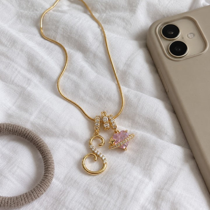 The gold Cherie initial necklace laid on a white duvet beside a phone, everyday phone photo