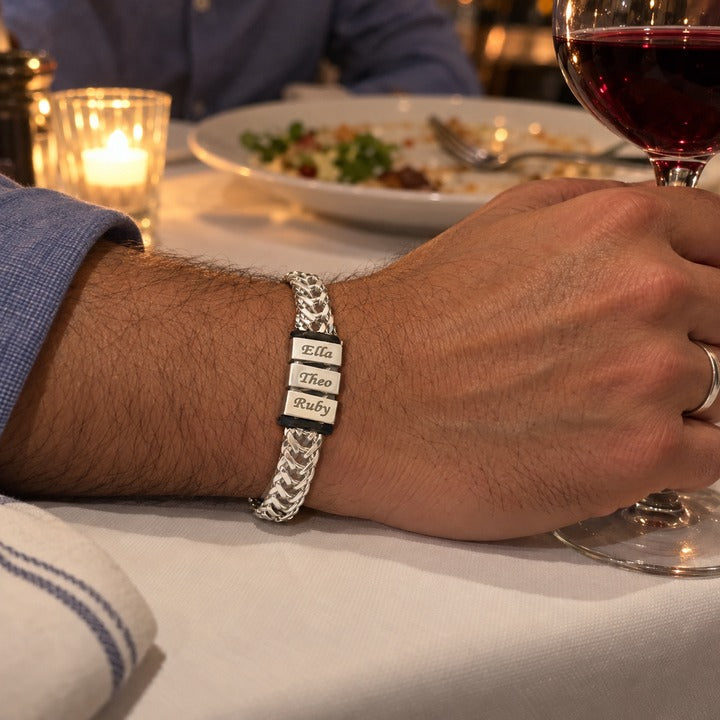 A man's forearm resting on a restaurant tablecloth at an anniversary dinner, his hand on a wine glass and the silver Harbor name bracelet with three names on his wrist, everyday phone photo
