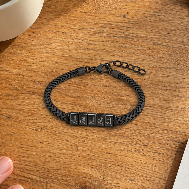 The black Harbor name bracelet with five names lying on a wooden kitchen table beside a mug, everyday phone photo