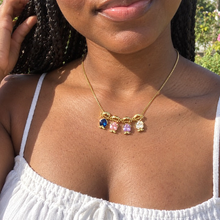 Phone photo of four charms on her collarbone in sunlight: royal blue, pale pink, pale lavender and clear heart-cut stones