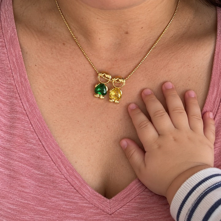 Looking down at her chest as a toddler's hand rests beside two boy charms with green and yellow heart-cut stones
